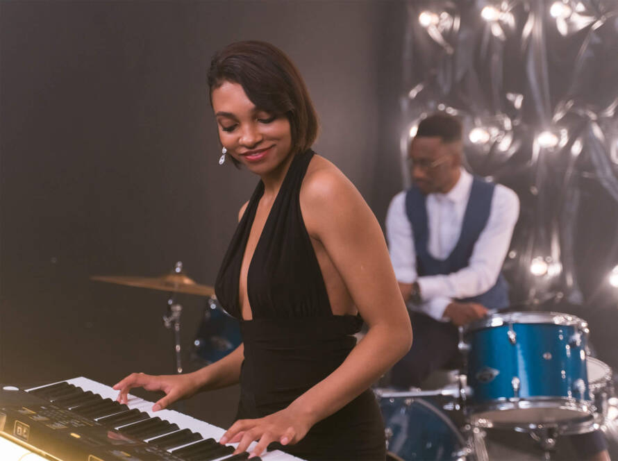 A woman in black dress playing piano while man plays drums.